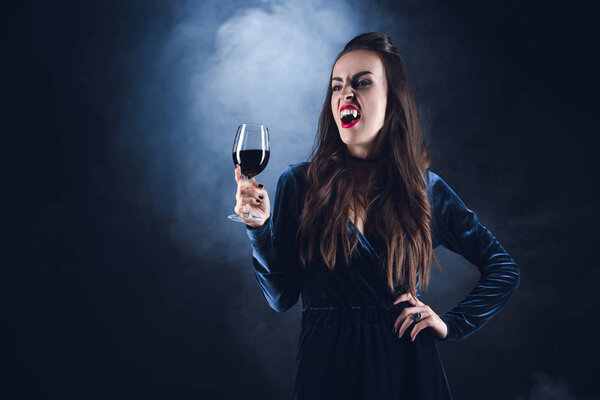 dreadful vampire holding wineglass with blood on dark background with smoke 