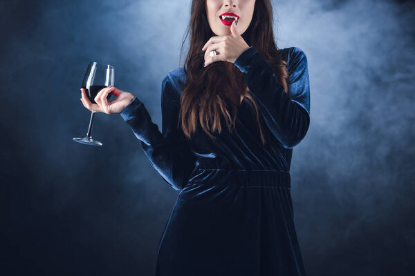 cropped view of vampire holding wineglass with blood and licking her fingers on dark background with smoke 