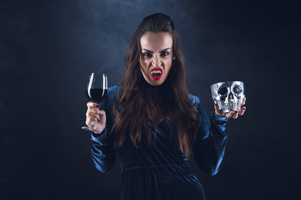 vampire holding metal skull and wineglass with blood on darkness with smoke 