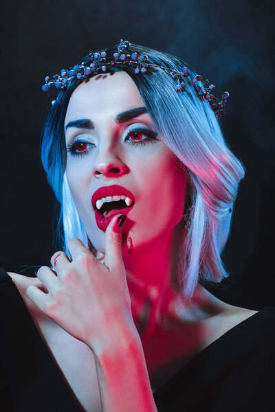 beautiful vampire woman touching her lips on dark background with smoke 
