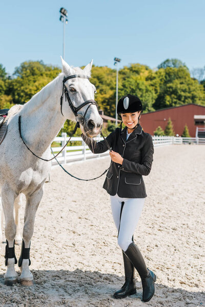 smiling attractive female equestrian looking at camera near horse at horse club