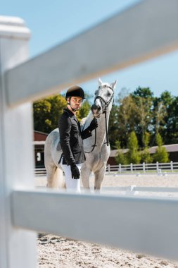 view through fence of handsome male equestrian standing near horse at horse club clipart