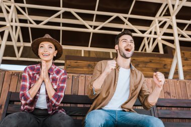 excited cowboy and cowgirl in casual clothes sitting on bench at ranch stadium and watching horse races clipart