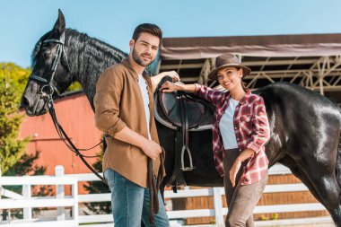 low angle view of cowboy and cowgirl standing near horse at ranch and looking at camera  clipart