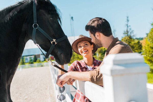 smiling cowboy and cowgirl standing near horse at ranch and looking at each other