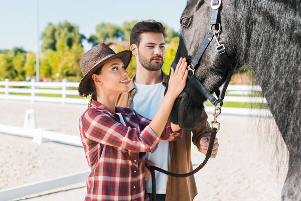 cowboy and cowgirl in casual clothes palming black horse at ranch