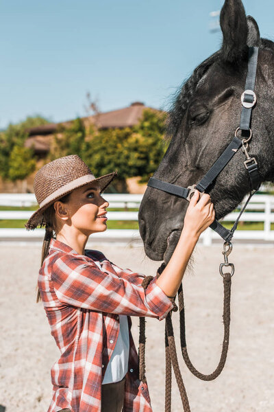 side view of attractive cowgirl in hat fixing horse halter at ranch