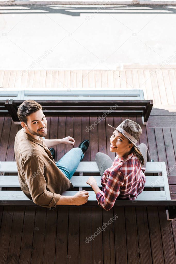 high angle view of smiling cowboy and cowgirl in casual clothes sitting on bench at ranch stadium