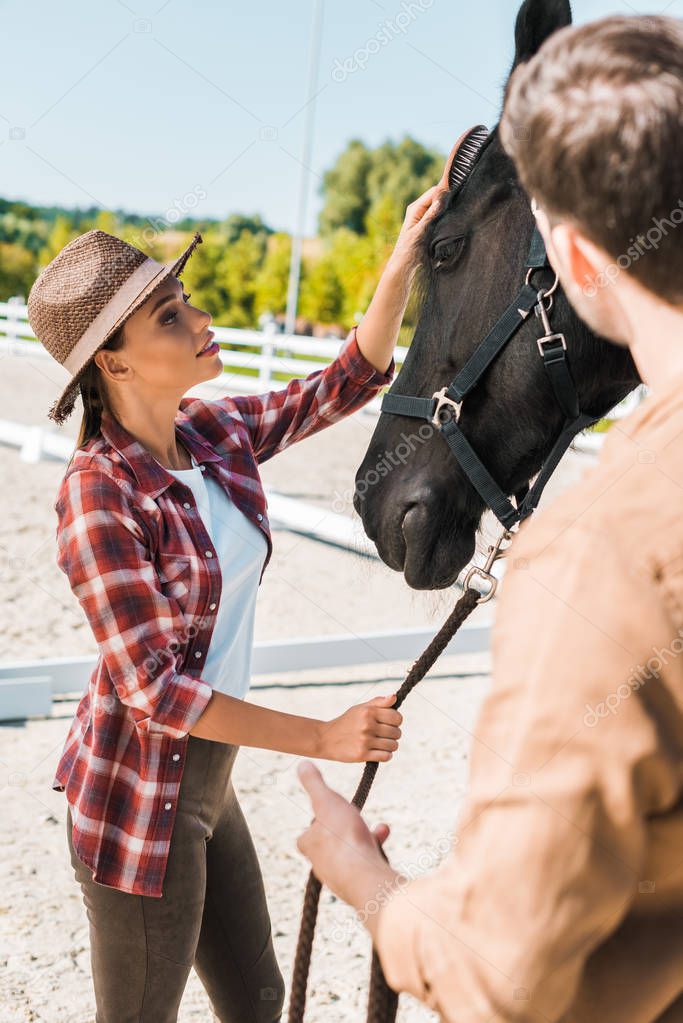 beautiful equestrian in hat palming black horse at ranch