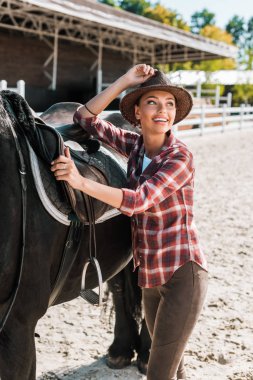 attractive smiling equestrian fixing horse saddle and touching hat at ranch clipart
