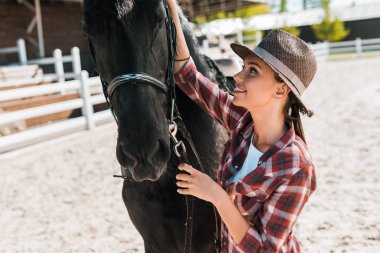attractive female equestrian in checkered shirt and hat fixing horse halter at ranch clipart