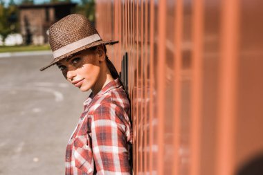 attractive cowgirl in checkered shirt and hat leaning on brown wall at ranch and looking at camera clipart