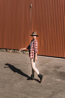 attractive cowgirl in checkered shirt and hat walking near brown building at ranch clipart