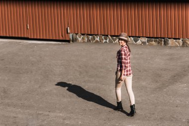 smiling attractive cowgirl in checkered shirt and hat walking near brown building at ranch clipart