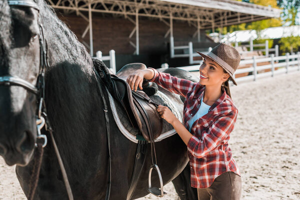attractive cowgirl in checkered shirt fixing horse saddle at ranch