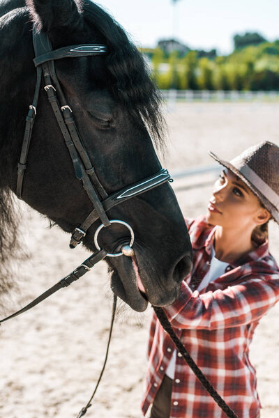 beautiful female equestrian in checkered shirt and hat fixing horse halter at ranch