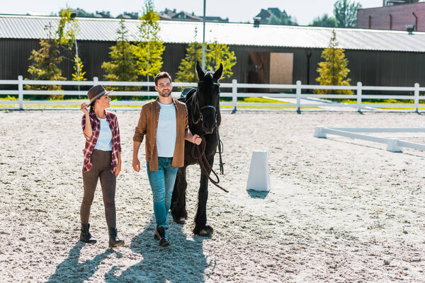 smiling cowboy and cowgirl in casual clothes walking with horse at ranch