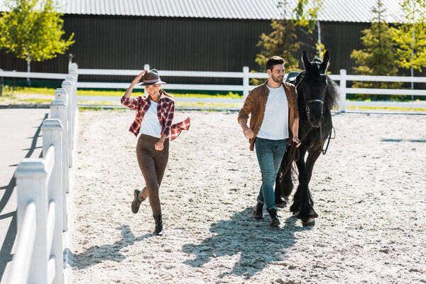 male equestrian walking with horse at ranch, happy female equestrian touching hat