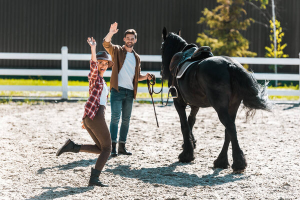 happy female and male equestrians walking with horse at ranch and waving hands