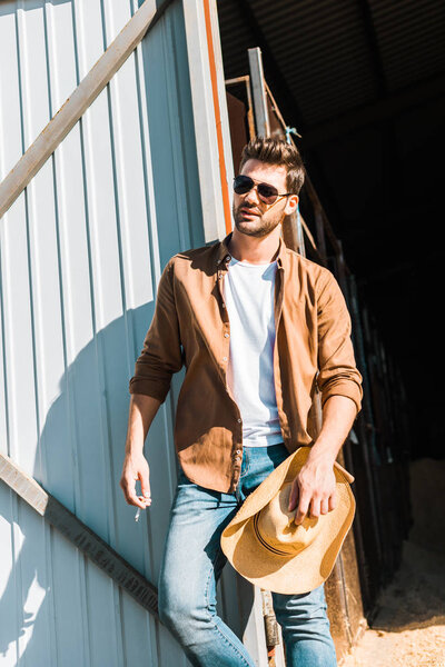 handsome man in sunglasses holding cigarette and hat, leaning on wall at ranch