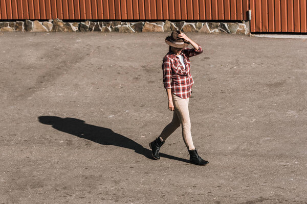 attractive woman in checkered shirt touching hat and walking near brown building at ranch