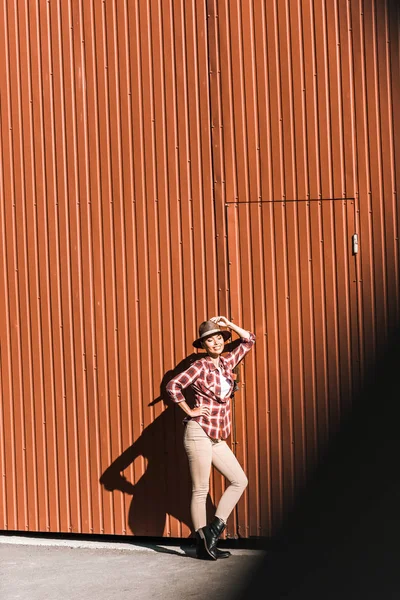 Улыбающаяся Привлекательная Ковбойша Клетчатой Рубашке Трогает Шляпу Опирается Коричневую Стену — Бесплатное стоковое фото