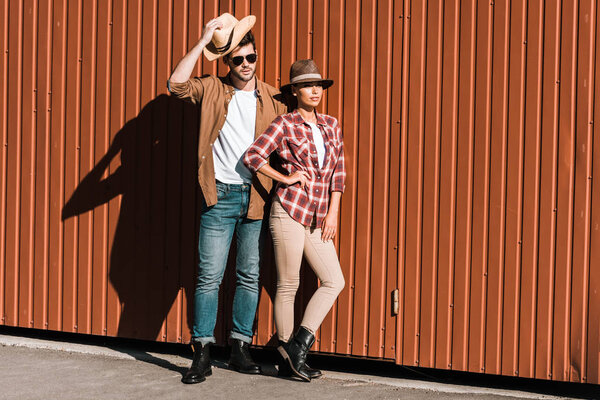 cowboy and cowgirl in casual clothes leaning on brown wall and looking away at ranch