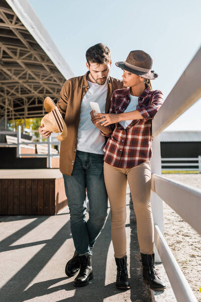 stylish equestrians in casual clothes looking at smartphone at ranch