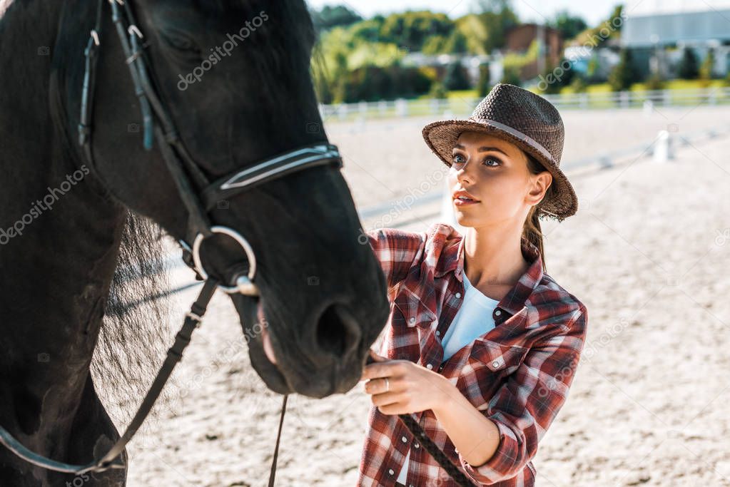 attractive jockey in checkered shirt and hat fixing horse halter at ranch