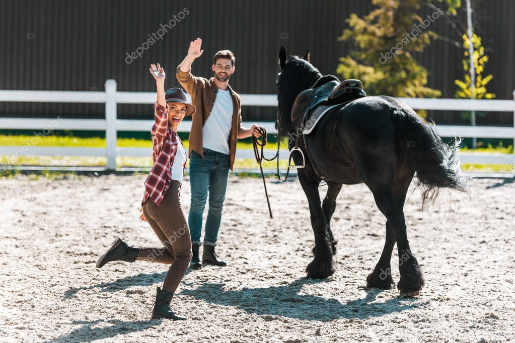 happy female and male equestrians walking with horse at ranch and waving hands