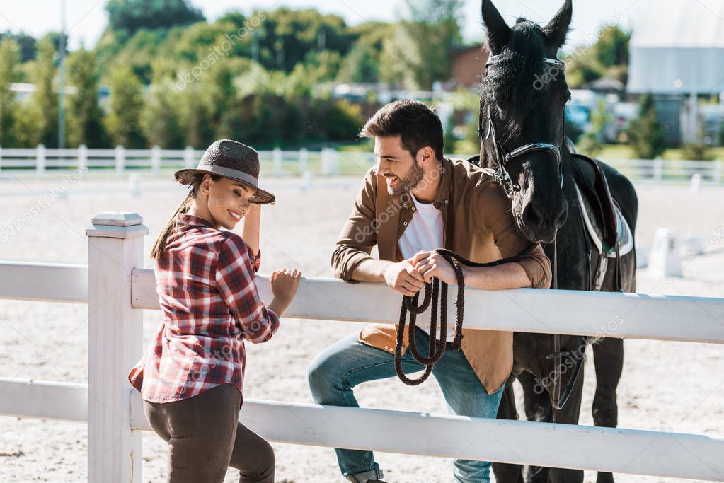 smiling cowboy and cowgirl standing near fence with horse and talking at ranch