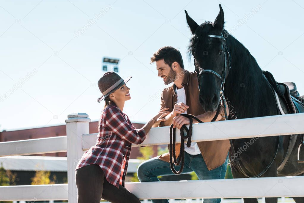 smiling female and male equestrians standing near fence with horse and looking at each other at ranch