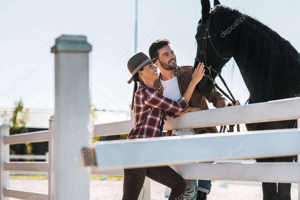 smiling cowboy and cowgirl standing near fence and palming horse at ranch