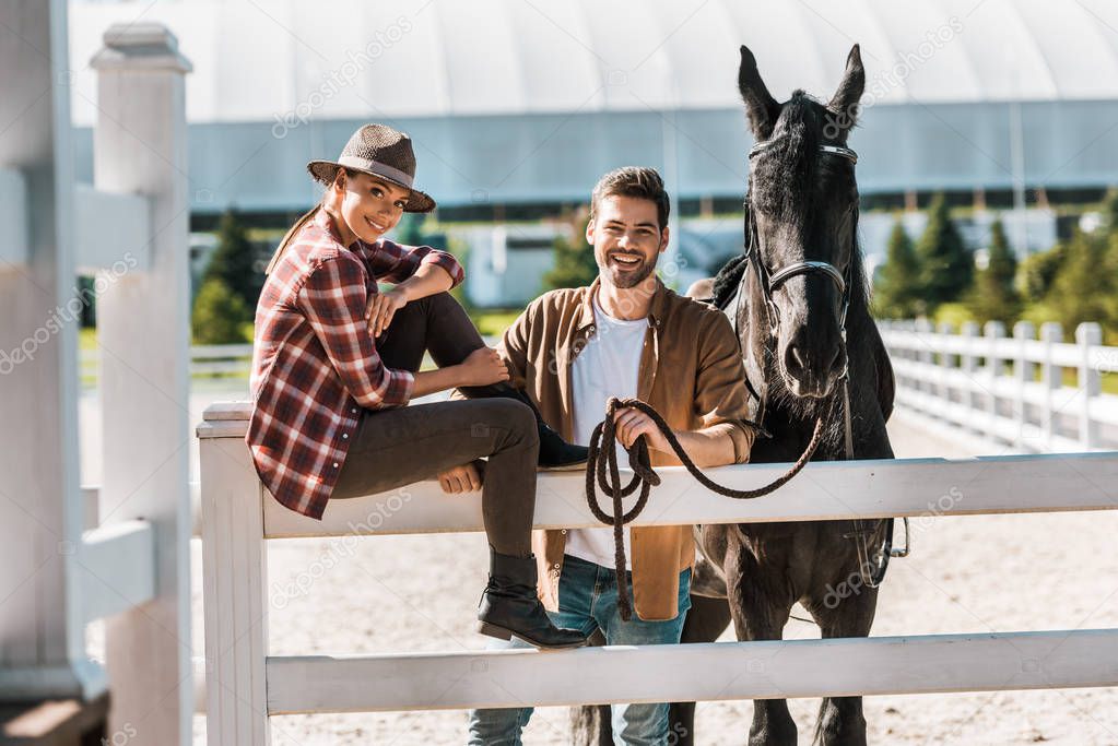 smiling female equestrian sitting on fence, colleague standing near fence with horse at ranch