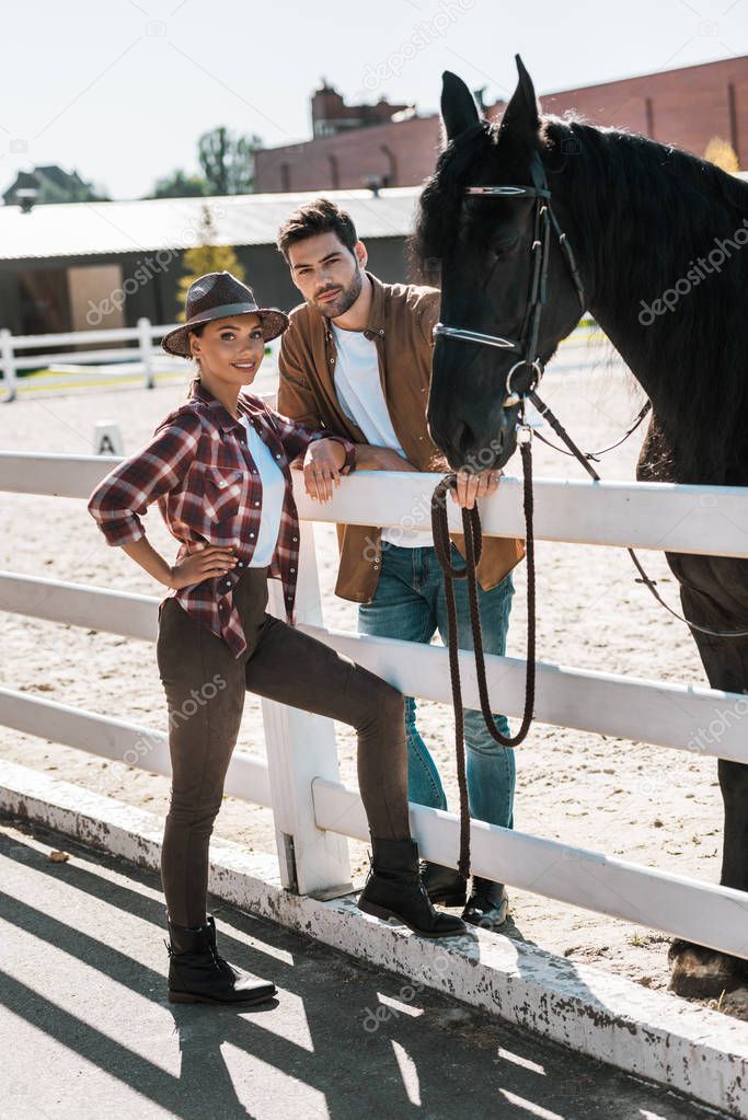 female and male equestrians standing near fence with horse at ranch and looking at camera