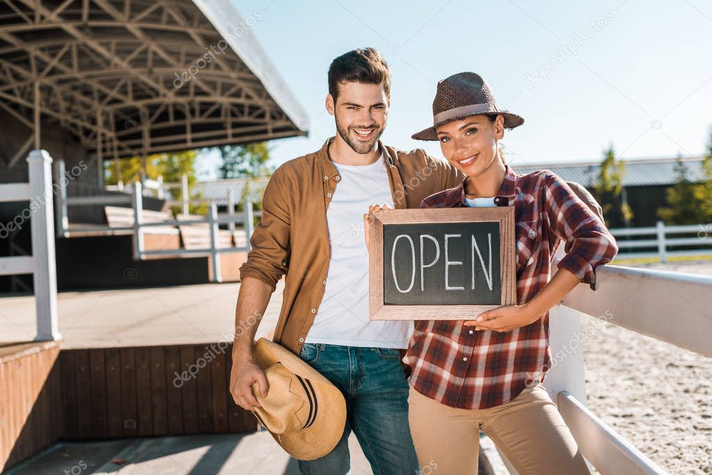 smiling stylish equestrians in casual clothes holding open sign at ranch and looking at camera