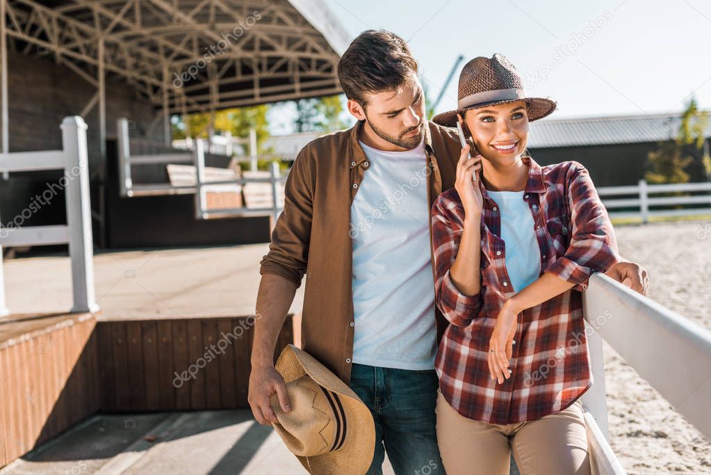 stylish cowboy and cowgirl in casual clothes standing near fence at ranch, woman talking by smartphone
