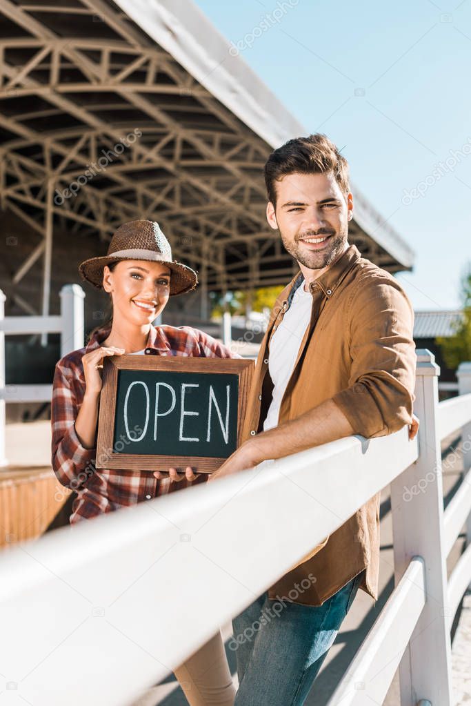 stylish cowboy and cowgirl in casual clothes standing near fence and holding open sign at ranch