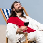 Selective focus of cheerful Jesus in robe and red sash resting on sun lounger with glass of red wine in desert