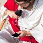 High angle view of Jesus resting on sun lounger, drinking wine and using smartphone with blank screen in desert