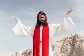 nízký úhel pohled na Ježíše v roucho, červenou šerpou a korunu z trní s zvedl ruce v poušti