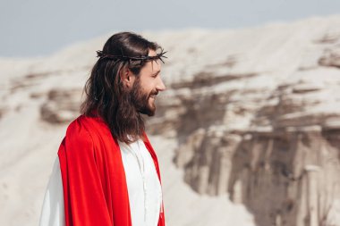 side view of smiling Jesus in robe, red sash and crown of thorns looking away in desert clipart