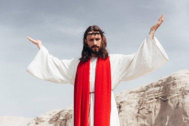 low angle view of Jesus in robe, red sash and crown of thorns standing with raised hands in desert