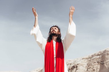 Jesus in robe, red sash and crown of thorns standing with raised hands and talking with god in desert clipart