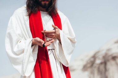 cropped image Jesus with long hair in robe and red sash holding wooden rosary in desert  clipart