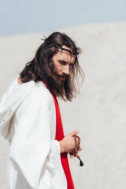 side view of Jesus in robe and red sash holding wooden rosary in desert  clipart