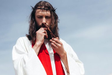 portrait of Jesus in robe, red sash and crown of thorns kissing rosary against blue sky clipart