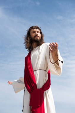 Jesus in robe, red sash and crown of thorns holding rosary and standing with open arms against blue sky clipart