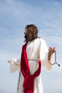 Jesus in robe, red sash and crown of thorns holding rosary and standing with open arms against blue sky, looking away clipart