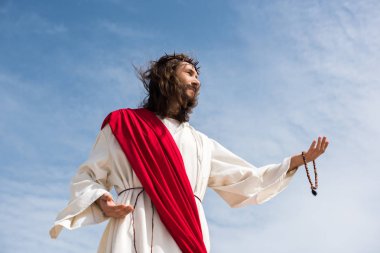 low angle view of Jesus in robe holding rosary and reaching hand against blue sky